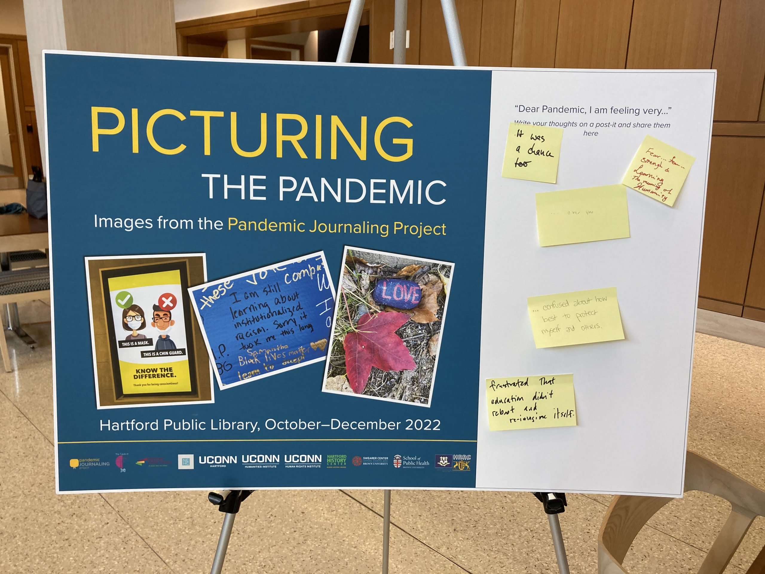A poster for the Picturing the Pandemic exhibit stands on an easel. The poster shows photographs taken by contributors to the pandemic journaling project and offers white space with the prompt "Dear Pandemic, I am feeling very..." A number of yellow post-it notes with writing on them have been affixed under the prompt.